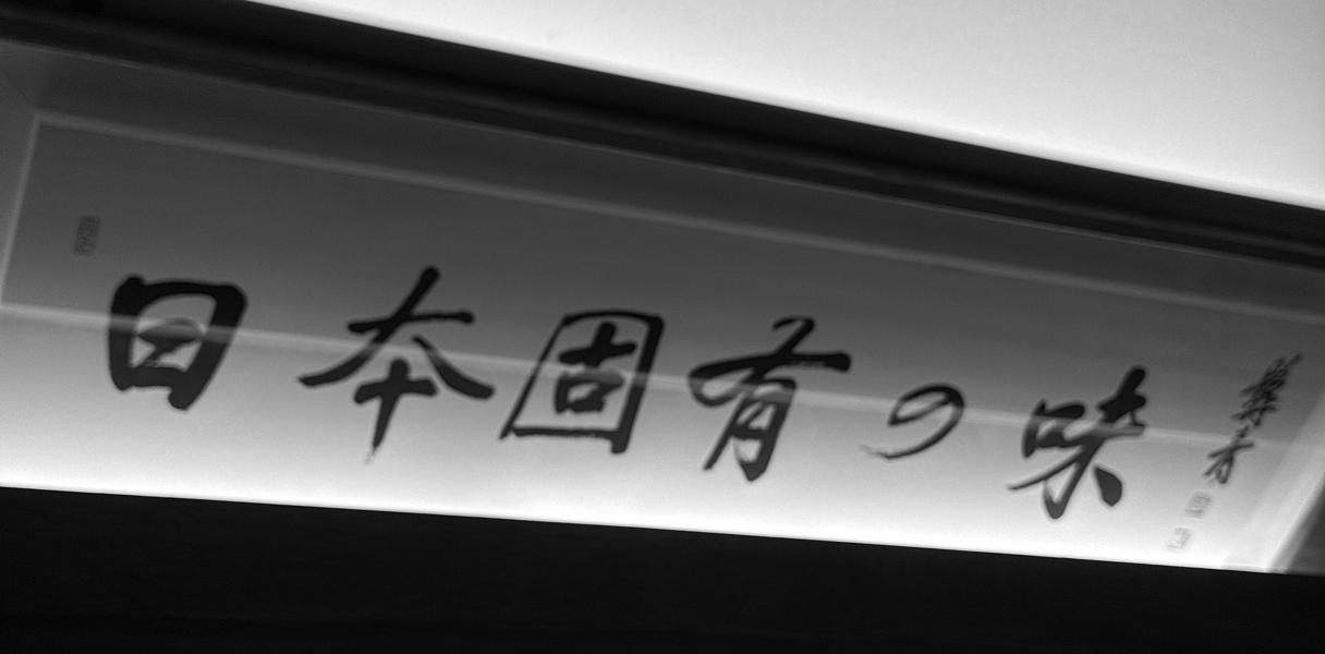 장 내부에 걸려 있는 '일본 고유의 맛(日本固有の味)'이라는 문구의 붓글씨 액자가 은은한 조명을 받아 정통 일식 선술집의 깊이 있는 분위기를 더해주는 모습